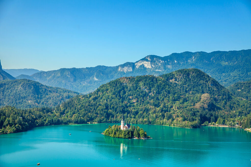 A picturesque view of Lake Bled with its iconic island and castle, surrounded by lush green mountains and clear blue skies.