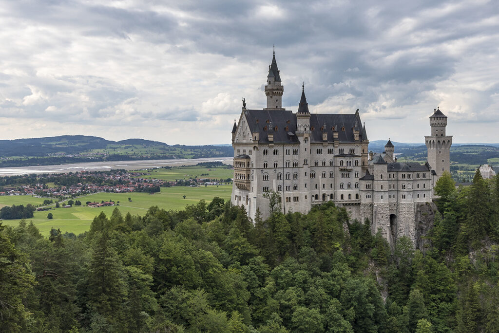 The Neuschwanstein Castle in Bavaria, nestled among the lush green hills, surrounded by forests and snow-capped mountains in the background.