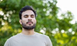 Young man engages in mindfulness and relaxation exercises, standing outdoors with eyes closed. Captures peaceful moments of meditation and wellness, highlighting focus on breathing